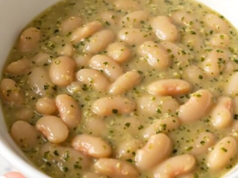 A top-down view of a slow cooker filled with white beans, chopped zucchini, and dollops of pesto and cream cheese.