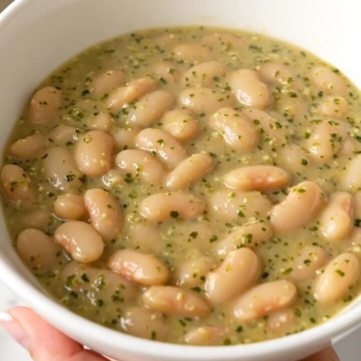 A top-down view of a slow cooker filled with white beans, chopped zucchini, and dollops of pesto and cream cheese.
