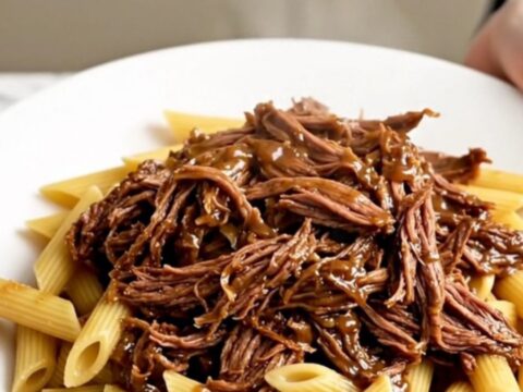 Slow cooker Tuscan shredded beef served over a bed of penne pasta on a white plate.