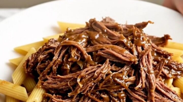 Slow cooker Tuscan shredded beef served over a bed of penne pasta on a white plate.