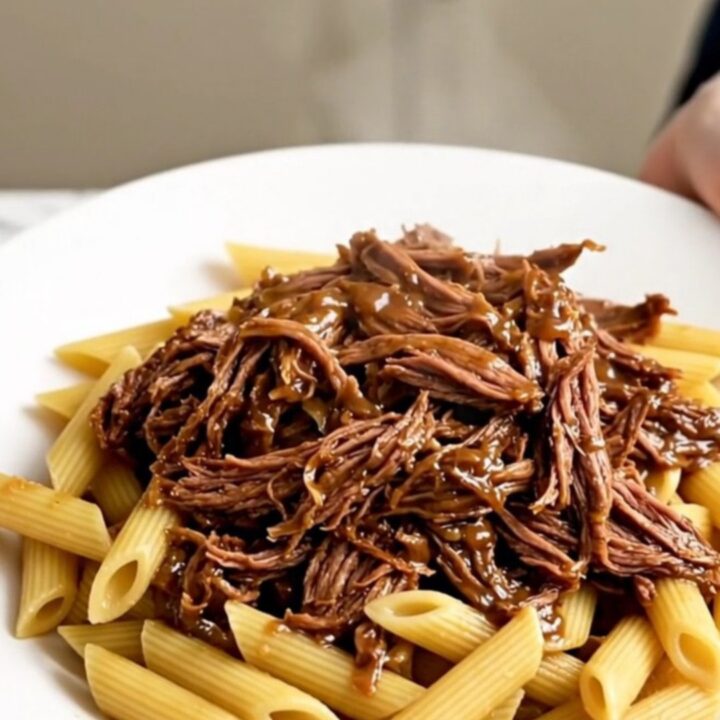 Slow cooker Tuscan shredded beef served over a bed of penne pasta on a white plate.