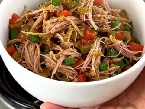 A close-up shot of a white bowl filled with slow-cooked shredded pork, black beans, and corn, topped with salsa verde.