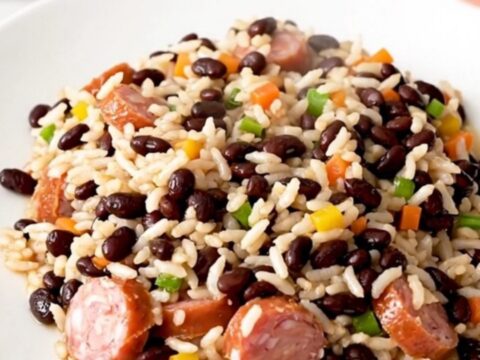 A close-up of a steaming, glossy dish of smoked sausage and black beans served on a white plate.