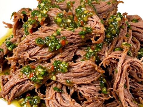 A close-up of tender, shredded beef in a slow cooker topped with vibrant green chimichurri sauce.
