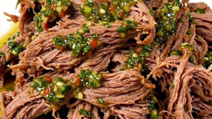 A close-up of tender, shredded beef in a slow cooker topped with vibrant green chimichurri sauce.