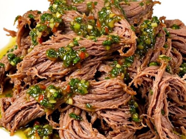 A close-up of tender, shredded beef in a slow cooker topped with vibrant green chimichurri sauce.