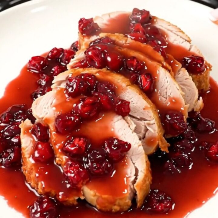 A close-up of tender, sliced turkey medallions coated in a glossy, vibrant red cranberry glaze on a white plate.
