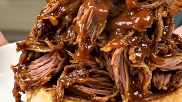 A close-up of a glossy, tender pulled pork sandwich on a sesame bun on a white plate.