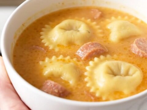 A steaming bowl of creamy ravioli and sausage soup on a white speckled countertop.