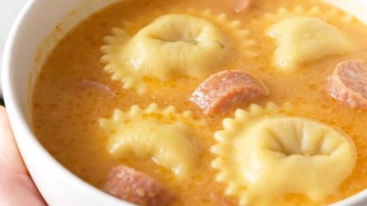 A steaming bowl of creamy ravioli and sausage soup on a white speckled countertop.