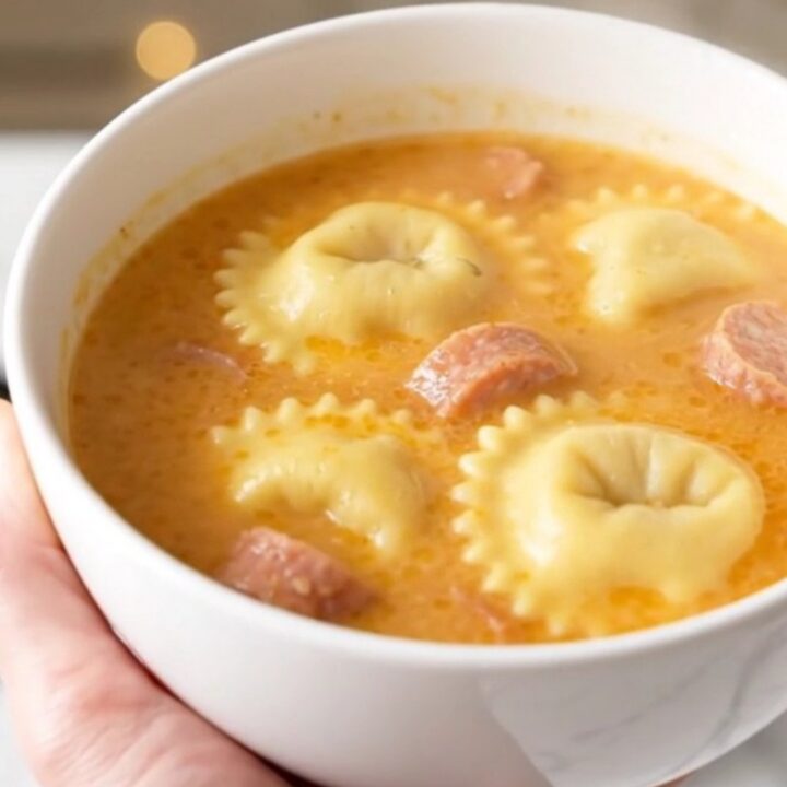 A steaming bowl of creamy ravioli and sausage soup on a white speckled countertop.