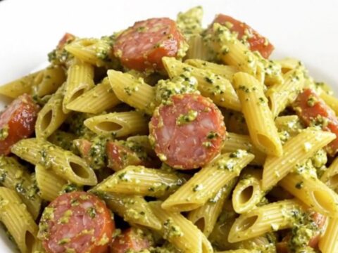 Creamy pesto Italian sausage penne served on a white plate with a rustic kitchen background.