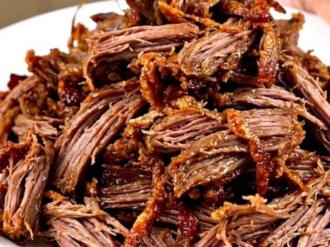 A close-up of tender, shredded beef carnitas on a white plate with fresh garnishes.