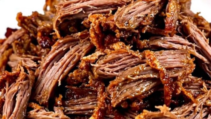 A close-up of tender, shredded beef carnitas on a white plate with fresh garnishes.