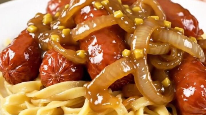 A close-up of glossy maple mustard glazed kielbasa and sliced onions served over buttery egg noodles on a white plate.