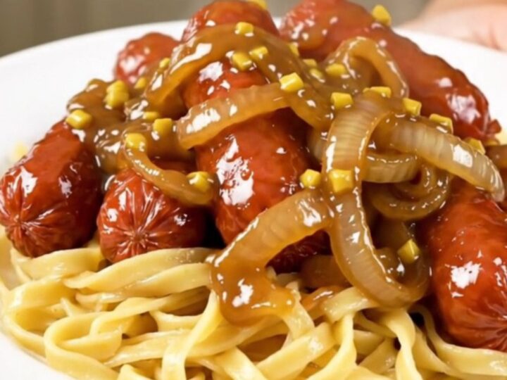 A close-up of glossy maple mustard glazed kielbasa and sliced onions served over buttery egg noodles on a white plate.