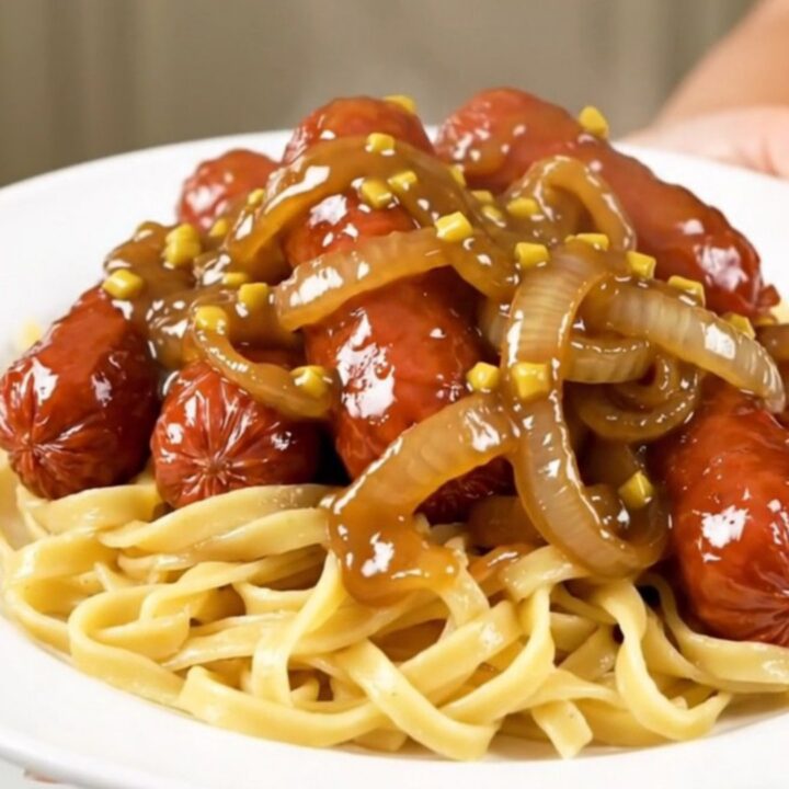 A close-up of glossy maple mustard glazed kielbasa and sliced onions served over buttery egg noodles on a white plate.
