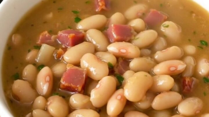 A rustic white bowl filled with creamy white bean and bacon stew, steaming hot and garnished with fresh herbs.