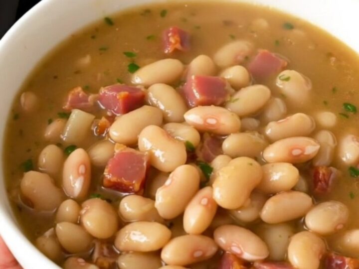A rustic white bowl filled with creamy white bean and bacon stew, steaming hot and garnished with fresh herbs.