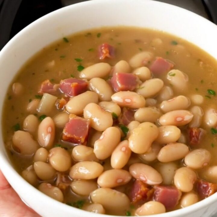 A rustic white bowl filled with creamy white bean and bacon stew, steaming hot and garnished with fresh herbs.