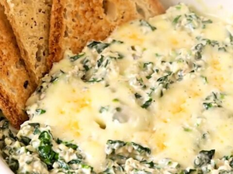 A top-down view of a black slow cooker filled with raw spinach, cream cheese, garlic, and shredded cheeses.