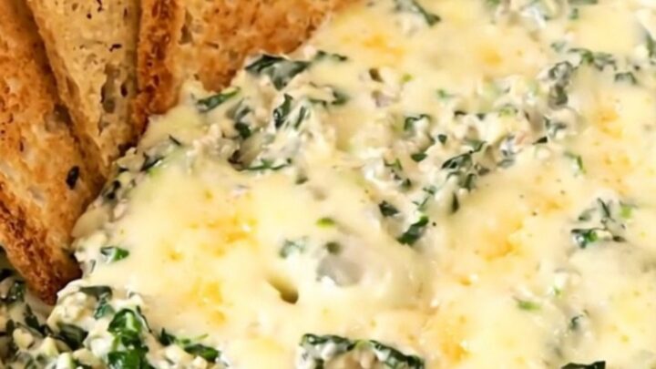 A top-down view of a black slow cooker filled with raw spinach, cream cheese, garlic, and shredded cheeses.