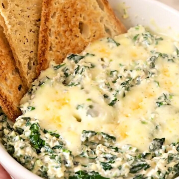 A top-down view of a black slow cooker filled with raw spinach, cream cheese, garlic, and shredded cheeses.