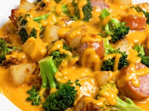 A close-up of a serving of cheesy broccoli, smoked sausage, and potatoes on a white plate.