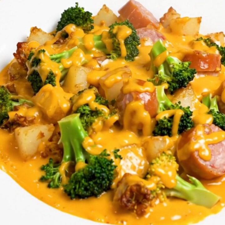 A close-up of a serving of cheesy broccoli, smoked sausage, and potatoes on a white plate.
