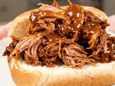 A close-up of a tender pulled pork sandwich on a soft roll served on a white plate.