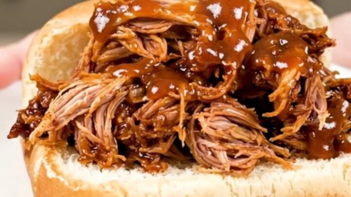 A close-up of a tender pulled pork sandwich on a soft roll served on a white plate.