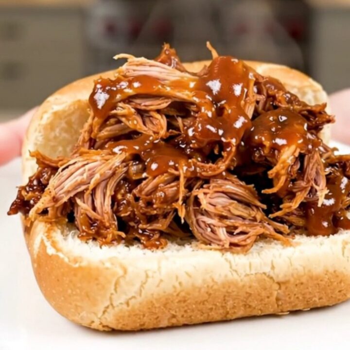 A close-up of a tender pulled pork sandwich on a soft roll served on a white plate.
