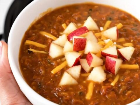 A bowl of hearty turkey chili topped with shredded white cheddar and diced apple against a blurred background.