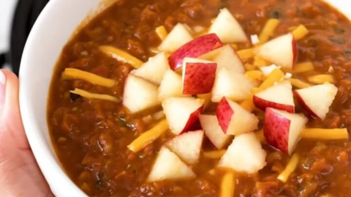A bowl of hearty turkey chili topped with shredded white cheddar and diced apple against a blurred background.