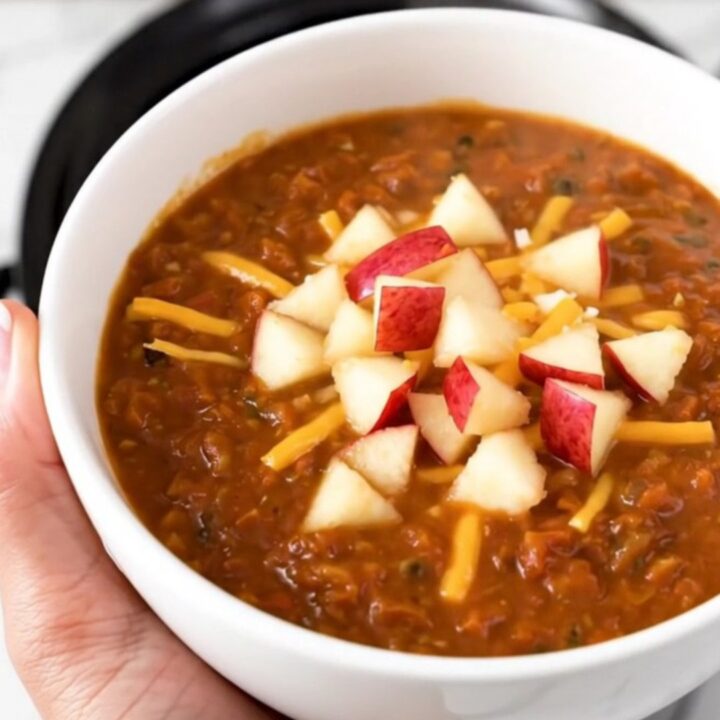 A bowl of hearty turkey chili topped with shredded white cheddar and diced apple against a blurred background.