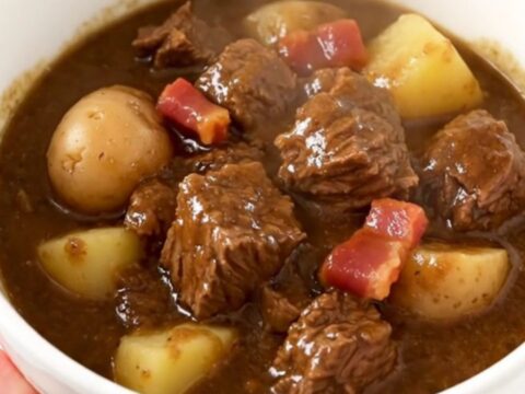 A warm bowl of rich beef stew with chunks of tender beef, potatoes, and bacon in a glossy savory sauce.