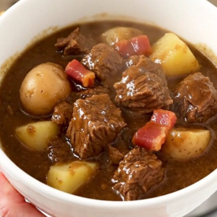 A warm bowl of rich beef stew with chunks of tender beef, potatoes, and bacon in a glossy savory sauce.