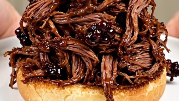 A close-up of glossy blackberry barbecue shredded beef piled high on a golden brioche bun.