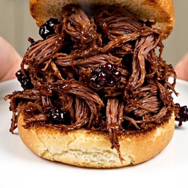 A close-up of glossy blackberry barbecue shredded beef piled high on a golden brioche bun.