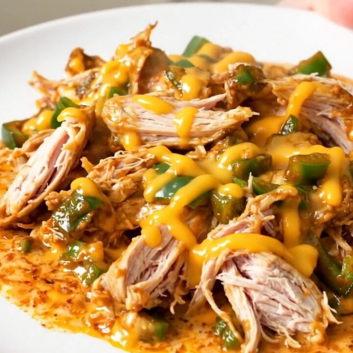 A top-down view of a black slow cooker filled with raw chicken breasts, diced poblano peppers, and a block of cream cheese.