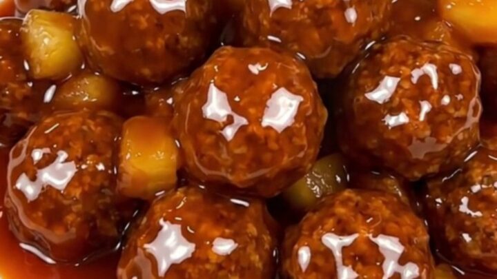 A top-down view of a slow cooker filled with beef meatballs, pineapple, and a thick red glaze.