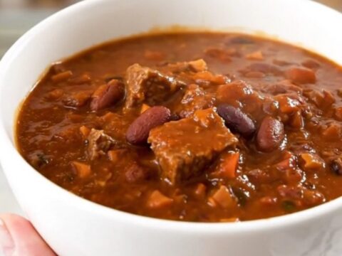 A white ceramic bowl filled with glossy, dark red beef chili, showing hearty chunks of beef and beans.