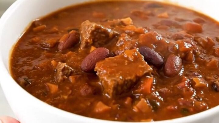 A white ceramic bowl filled with glossy, dark red beef chili, showing hearty chunks of beef and beans.