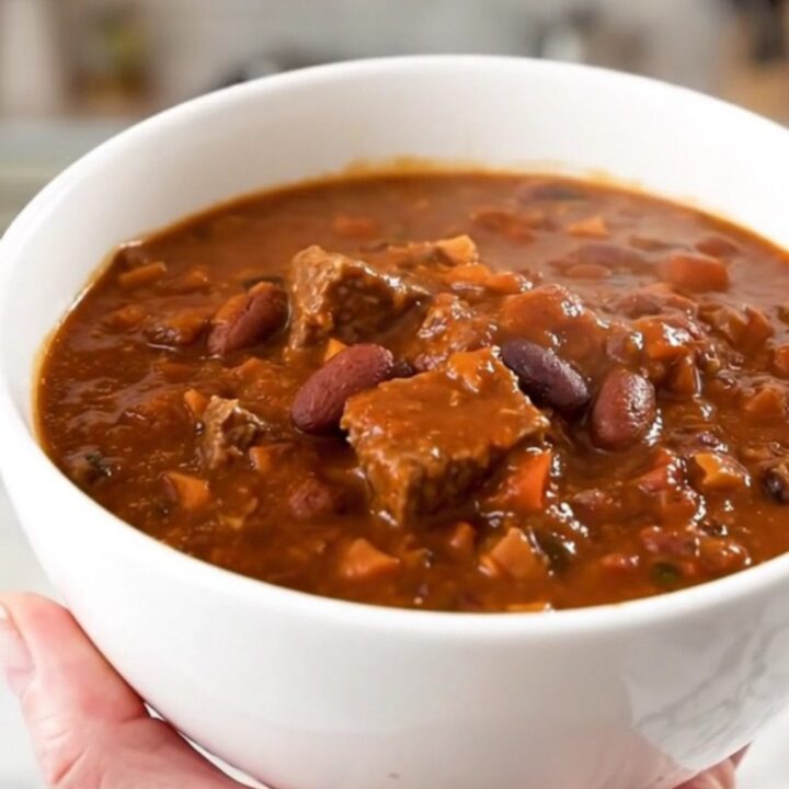 A white ceramic bowl filled with glossy, dark red beef chili, showing hearty chunks of beef and beans.