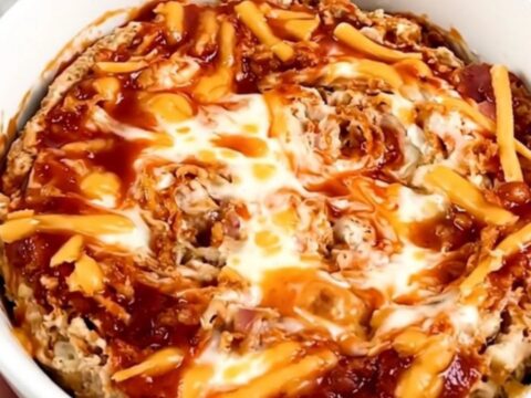 A top-down view of a black slow cooker filled with layers of cream cheese, bacon, shredded cheese, and buffalo sauce.