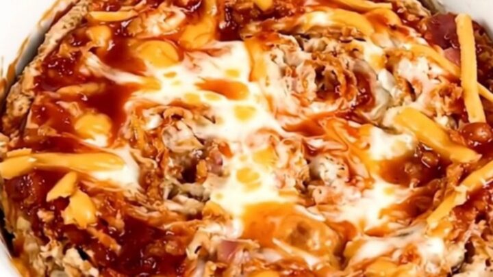 A top-down view of a black slow cooker filled with layers of cream cheese, bacon, shredded cheese, and buffalo sauce.