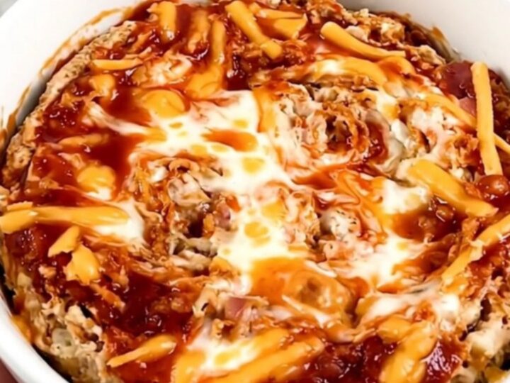 A top-down view of a black slow cooker filled with layers of cream cheese, bacon, shredded cheese, and buffalo sauce.