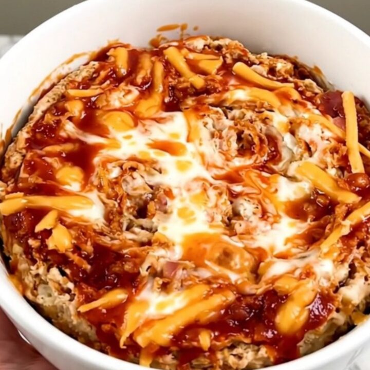 A top-down view of a black slow cooker filled with layers of cream cheese, bacon, shredded cheese, and buffalo sauce.