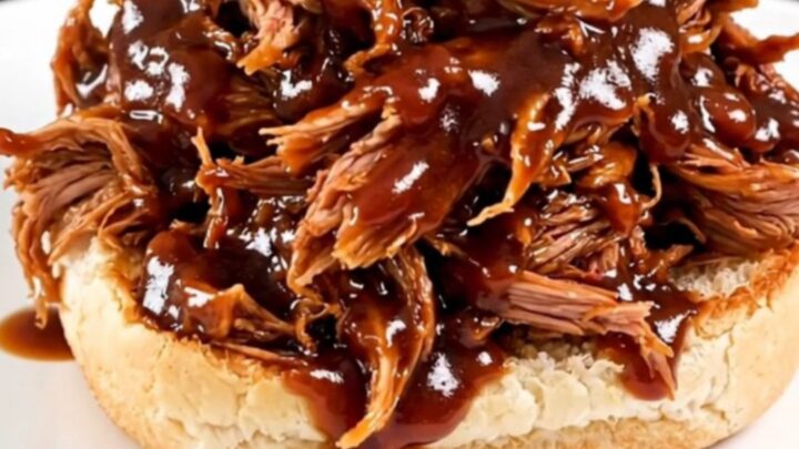 A close-up of glossy, shredded pulled pork piled high on a white bun on a plain round plate.