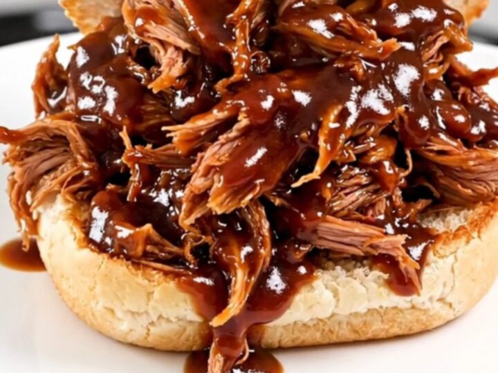 A close-up of glossy, shredded pulled pork piled high on a white bun on a plain round plate.